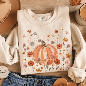 May include: A sand-colored crewneck sweatshirt featuring a central graphic of a large orange pumpkin surrounded by a variety of painted-style wildflowers in shades of red, orange, white, and muted blue, with long sleeves and a ribbed collar.