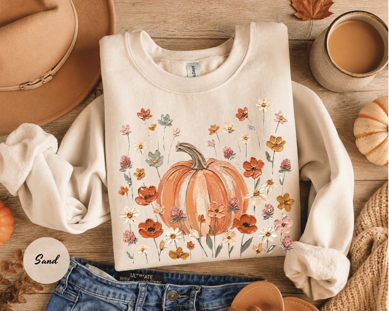 May include: A sand-colored crewneck sweatshirt featuring a central graphic of a large orange pumpkin surrounded by a variety of painted-style wildflowers in shades of red, orange, white, and muted blue, with long sleeves and a ribbed collar.