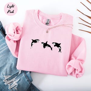 May include: Light pink sweatshirt with three black and white orca whale graphics. The sweatshirt has a crew neck and long sleeves. The words "Light Pink" are in a pink circle in the upper left corner.