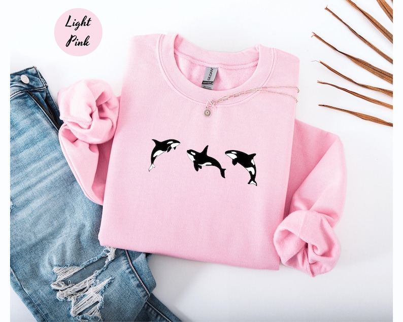 May include: Light pink sweatshirt with three black and white orca whale graphics. The sweatshirt has a crew neck and long sleeves. The words "Light Pink" are in a pink circle in the upper left corner.