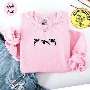 May include: Light pink sweatshirt with three black orca whale designs. The sweatshirt is paired with blue jeans. The text "Light Pink" is in a pink circle in the upper left corner. The word "Embroidery" is in a yellow circle in the upper right corner.