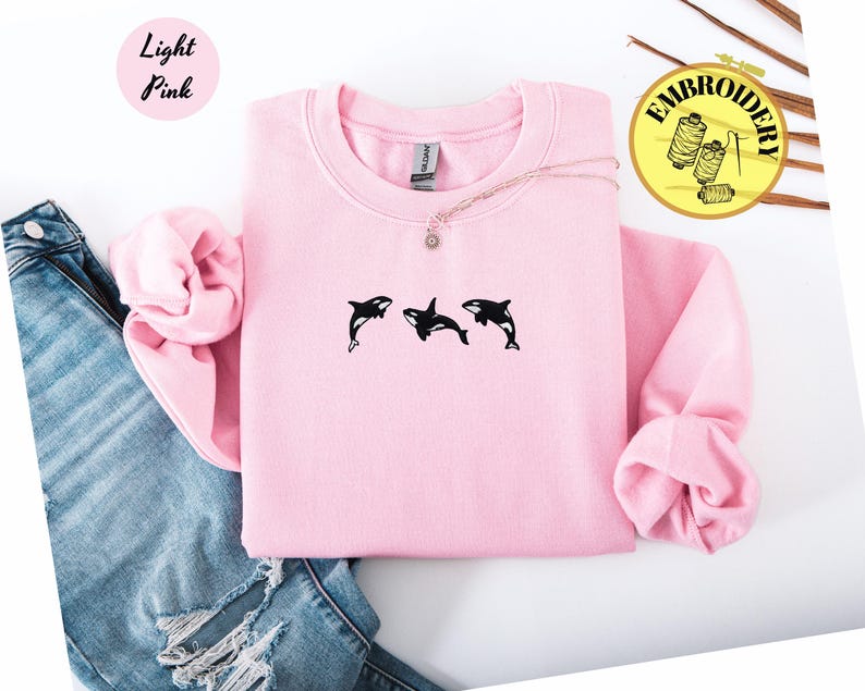 May include: Light pink sweatshirt with three black orca whale designs. The sweatshirt is paired with blue jeans. The text "Light Pink" is in a pink circle in the upper left corner. The word "Embroidery" is in a yellow circle in the upper right corner.