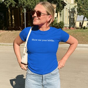 May include: A woman in a royal blue t-shirt with the phrase "Show me your books." The shirt is worn with light-wash denim jeans and a white shoulder bag. She is wearing sunglasses, standing outdoors on a sunny day.