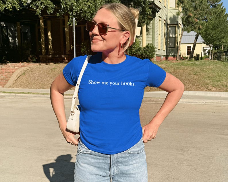 May include: A woman in a royal blue t-shirt with the phrase "Show me your books." The shirt is worn with light-wash denim jeans and a white shoulder bag. She is wearing sunglasses, standing outdoors on a sunny day.