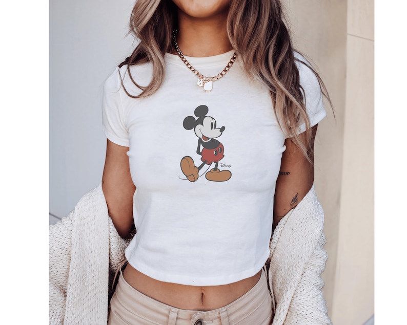 Może przedstawiać: Biały crop top z grafiką Myszki Miki. Postać z kresk&oacute;wki jest przedstawiona w kolorach czarnym, czerwonym i beżowym. Koszulka ma kr&oacute;tkie rękawy i okrągły dekolt, idealna do noszenia na co dzień. Widoczne logo Disneya.