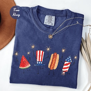 K&ouml;nnte beinhalten: Marineblaues T-Shirt mit einer skurrilen Illustration einer Wassermelonenscheibe, eines Hutes, eines Hotdogs und eines Feuerwerks, die jeweils Wunderkerzen halten. Das Shirt hat den Text "True Navy" und ein Comfort Colors-Etikett.