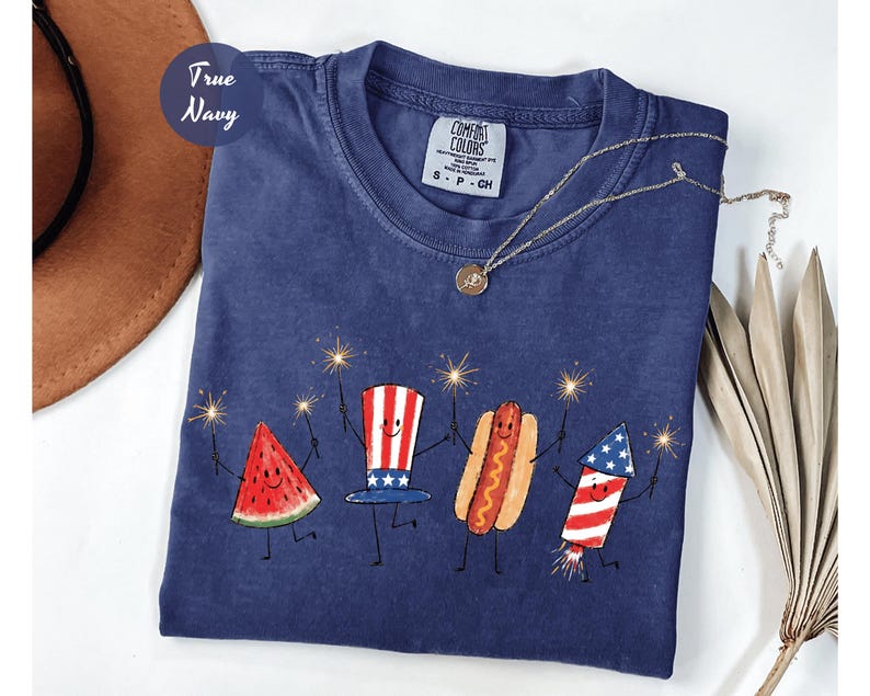 K&ouml;nnte beinhalten: Marineblaues T-Shirt mit einer skurrilen Illustration einer Wassermelonenscheibe, eines Hutes, eines Hotdogs und eines Feuerwerks, die jeweils Wunderkerzen halten. Das Shirt hat den Text "True Navy" und ein Comfort Colors-Etikett.