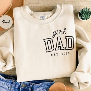 May include: A cream-colored sweatshirt with the words "girl DAD EST. 2025" printed in black. The sweatshirt is folded on a wooden surface, with a pair of blue jeans and a hat in the background. A small succulent plant is also visible.