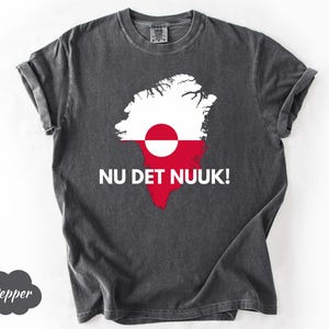 May include: Dark gray t-shirt featuring a graphic of the Greenland flag in red, white, and a red circle. The text "NU DET NUUK!" is printed below the graphic. The shirt has a crew neck and short sleeves.