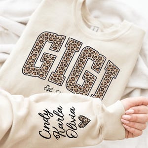 May include: Cream-colored sweatshirt with the word "GIGI" in a leopard print design. The sleeve features the names "Cindy, Karla, Olivia" in black script. A heart graphic is also on the sleeve. The sweatshirt is folded on a white surface.