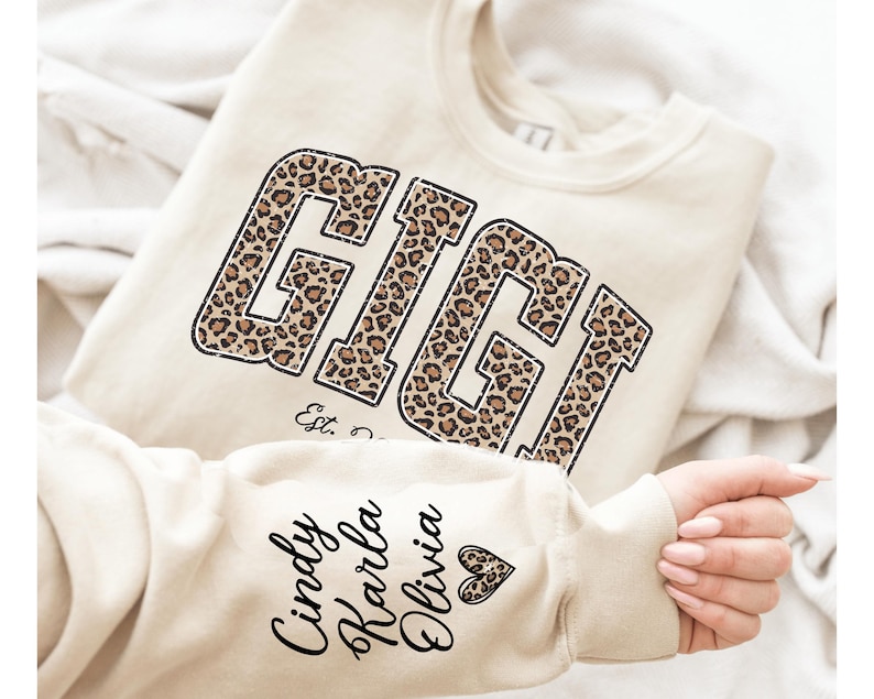 May include: Cream-colored sweatshirt with the word "GIGI" in a leopard print design. The sleeve features the names "Cindy, Karla, Olivia" in black script. A heart graphic is also on the sleeve. The sweatshirt is folded on a white surface.