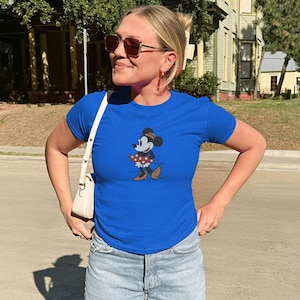 Pu&ograve; includere: Una maglietta blu reale con una grafica di Minnie Mouse. Il personaggio dei cartoni animati &egrave; raffigurato in una posa classica, con una gonna a pois e scarpe marroni. La persona indossa occhiali da sole e una borsa bianca.