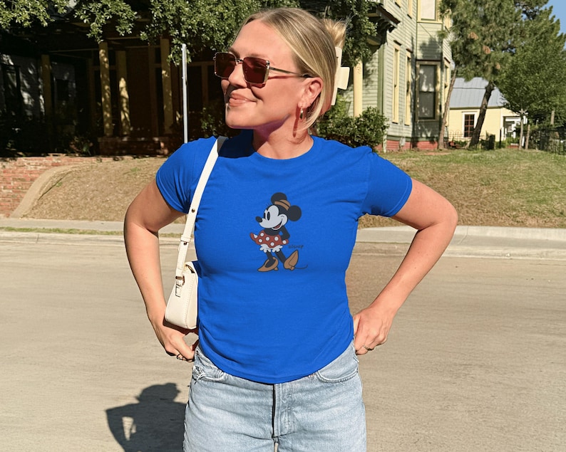 Pu&ograve; includere: Una maglietta blu reale con una grafica di Minnie Mouse. Il personaggio dei cartoni animati &egrave; raffigurato in una posa classica, con una gonna a pois e scarpe marroni. La persona indossa occhiali da sole e una borsa bianca.