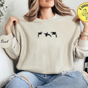 May include: A sand-colored sweatshirt featuring a black embroidered design of three orcas. The sweatshirt has a crew neck and long sleeves. The word "Sand" is printed on a round sticker.
