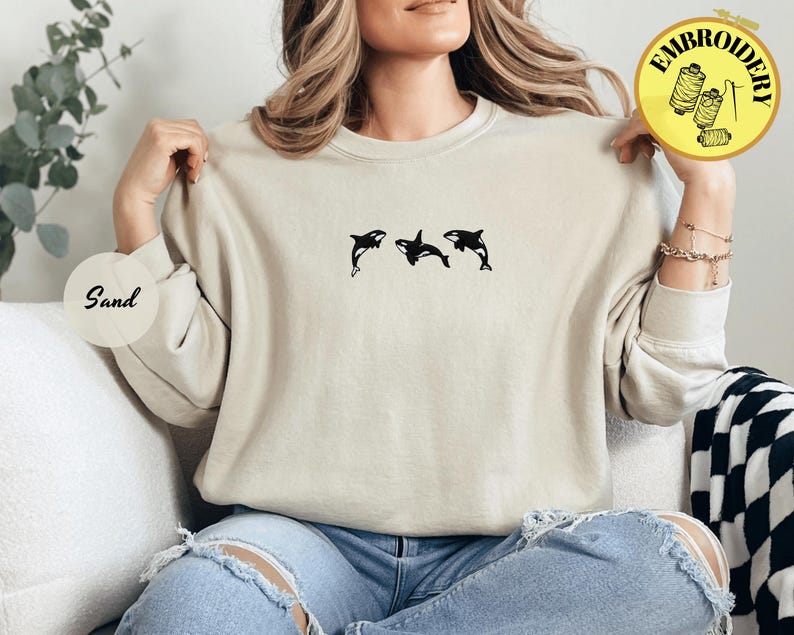 May include: A sand-colored sweatshirt featuring a black embroidered design of three orcas. The sweatshirt has a crew neck and long sleeves. The word "Sand" is printed on a round sticker.