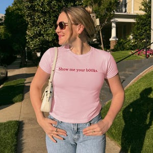 May include: A pale pink t-shirt with the phrase "Show me your books" in red. The top is paired with blue jeans. A white handbag is worn over the shoulder.
