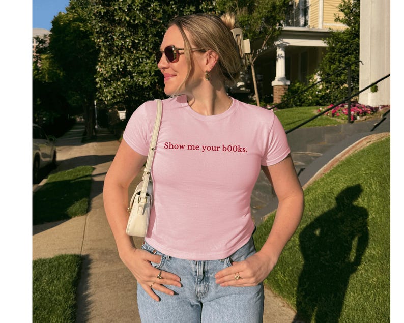 May include: A pale pink t-shirt with the phrase "Show me your books" in red. The top is paired with blue jeans. A white handbag is worn over the shoulder.