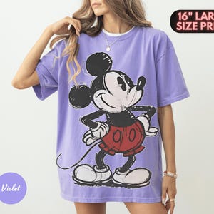 Puede incluir: Una camiseta violeta con un gran gr&aacute;fico de Mickey Mouse en negro, blanco y rojo. La camiseta tiene mangas cortas y un corte holgado. El texto en la camiseta dice "16" LARGE SIZE PRINT" y "Violet".