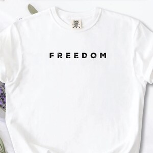 May include: A white t-shirt with the word "FREEDOM" printed in black, centered on the chest. The shirt has short sleeves and a crew neck. The background includes lavender sprigs and a white surface.