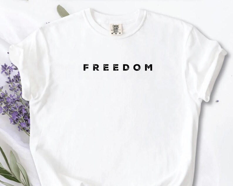 May include: A white t-shirt with the word "FREEDOM" printed in black, centered on the chest. The shirt has short sleeves and a crew neck. The background includes lavender sprigs and a white surface.