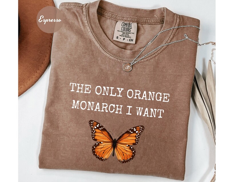 Pode incluir: Camiseta cor espresso com o texto "THE ONLY ORANGE MONARCH I WANT" e um gr&aacute;fico de borboleta monarca. Um colar de prata com um pequeno pingente repousa sobre a camisa. A camisa tem uma etiqueta Comfort Colors.