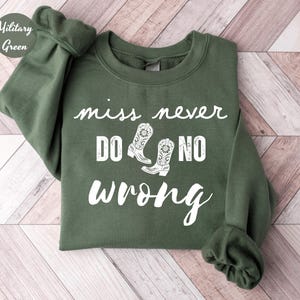 May include: Military green sweatshirt with the text "miss never do no wrong" in white script. The design includes white cowboy boot illustrations. The sweatshirt is folded on a light wood grain surface.
