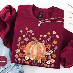 May include: A maroon crewneck sweatshirt featuring a central graphic of a large orange pumpkin surrounded by a variety of colorful wildflowers in shades of yellow, orange, and pink. The garment has long sleeves and a ribbed collar and cuffs.