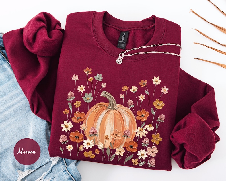 May include: A maroon crewneck sweatshirt featuring a central graphic of a large orange pumpkin surrounded by a variety of colorful wildflowers in shades of yellow, orange, and pink. The garment has long sleeves and a ribbed collar and cuffs.