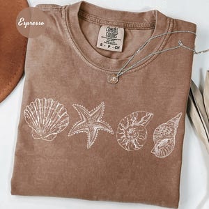 May include: Espresso-colored t-shirt with a crew neck and white seashell graphics. The shirt features a starfish, scallop shell, nautilus shell, and conch shell design. A silver necklace with a small pendant rests on the shirt. The tag reads "Comfort Colors".