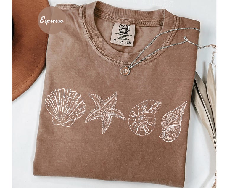 May include: Espresso-colored t-shirt with a crew neck and white seashell graphics. The shirt features a starfish, scallop shell, nautilus shell, and conch shell design. A silver necklace with a small pendant rests on the shirt. The tag reads "Comfort Colors".