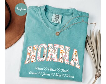 May include: A teal blue t-shirt with the word "NONNA" in white letters outlined with floral designs. Below the word are the names Liam, Olivia, Noah, Emma, James, Ava, and Lucas with a heart symbol between each name.