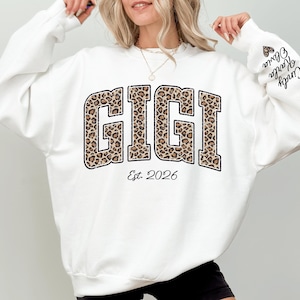May include: A white sweatshirt with the word "GIGI" in a leopard print design. The text "Est. 2026" is below the word "GIGI". The sleeve has the names "Cindy Karla Olivia" printed on it.