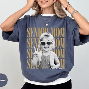 May include: A denim-coloured short-sleeve t-shirt featuring a black and white photograph of a child wearing heart-shaped sunglasses and making a peace sign. The text SENIOR MOM is repeated in gold behind the photo, with the name Amelia in script.