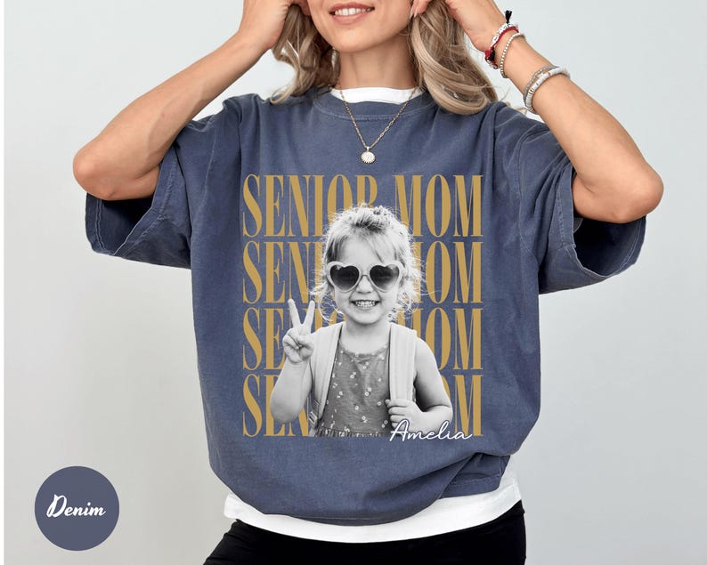 May include: A denim-coloured short-sleeve t-shirt featuring a black and white photograph of a child wearing heart-shaped sunglasses and making a peace sign. The text SENIOR MOM is repeated in gold behind the photo, with the name Amelia in script.