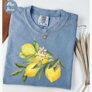 May include: A light blue Comfort Colors t-shirt featuring a lemon branch design with yellow lemons, green leaves, and white blossoms. The shirt has a crew neck and short sleeves. A silver necklace with a pendant is draped on the shirt. The words "Blue Jean" are in the upper left corner.