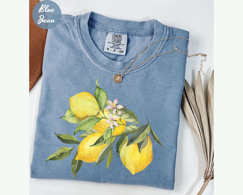 May include: A light blue Comfort Colors t-shirt featuring a lemon branch design with yellow lemons, green leaves, and white blossoms. The shirt has a crew neck and short sleeves. A silver necklace with a pendant is draped on the shirt. The words "Blue Jean" are in the upper left corner.