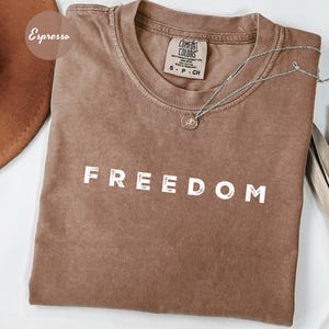 May include: Espresso-colored t-shirt with the word "FREEDOM" printed in white block letters. The shirt has a crew neck and short sleeves. A silver necklace with a small pendant rests on the shirt.