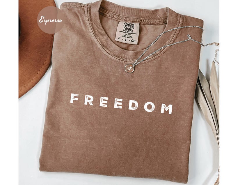 May include: Espresso-colored t-shirt with the word "FREEDOM" printed in white block letters. The shirt has a crew neck and short sleeves. A silver necklace with a small pendant rests on the shirt.