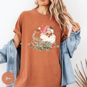 May include: A rust-colored t-shirt featuring a vintage illustration of Santa Claus with a reindeer and festive greenery. Santa wears a pink hat and has a long white beard. The design evokes a classic Christmas theme.