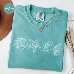 May include: Seafoam green t-shirt with white seashell graphics. The shirt features a crew neck and short sleeves. A gold necklace with a pendant rests on the shirt. The word "Seafoam" is in a circle in the upper left corner.