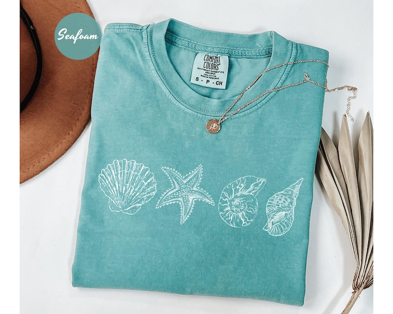 May include: Seafoam green t-shirt with white seashell graphics. The shirt features a crew neck and short sleeves. A gold necklace with a pendant rests on the shirt. The word "Seafoam" is in a circle in the upper left corner.