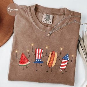 K&ouml;nnte beinhalten: Espressofarbenes T-Shirt mit einem skurrilen Design von animierten Icons zum 4. Juli, die Wunderkerzen halten. Das Design umfasst eine Wassermelonenscheibe, einen Uncle-Sam-Hut, einen Hotdog und ein Feuerwerk. Eine silberne Halskette liegt auf dem Shirt.