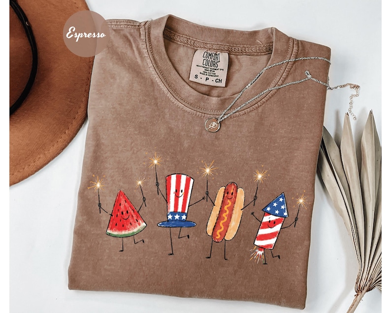 K&ouml;nnte beinhalten: Espressofarbenes T-Shirt mit einem skurrilen Design von animierten Icons zum 4. Juli, die Wunderkerzen halten. Das Design umfasst eine Wassermelonenscheibe, einen Uncle-Sam-Hut, einen Hotdog und ein Feuerwerk. Eine silberne Halskette liegt auf dem Shirt.