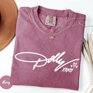 May include: A berry-colored short-sleeve t-shirt featuring the white script text Dolly and xoxo with three small heart outlines. The shirt has a crew neckline and a Comfort Colors brand label on the inside collar with size S-CH indicated.