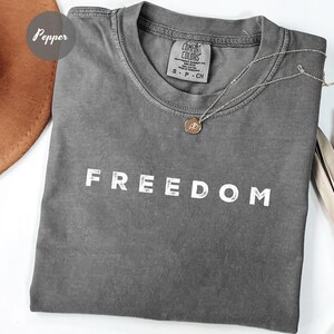 May include: A gray Comfort Colors t-shirt with the word "FREEDOM" printed in white. A silver necklace with a pendant is draped across the shirt. The shirt is folded and has a small tag.