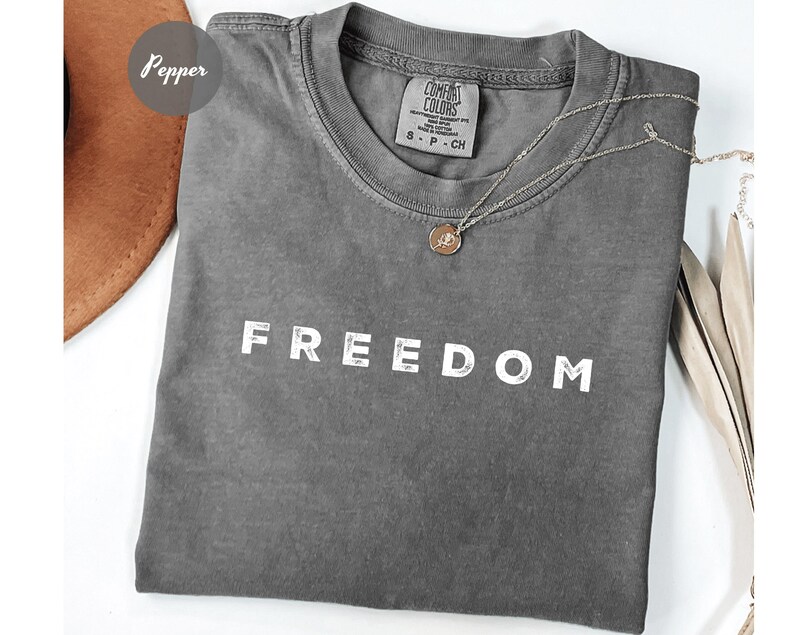May include: A gray Comfort Colors t-shirt with the word "FREEDOM" printed in white. A silver necklace with a pendant is draped across the shirt. The shirt is folded and has a small tag.