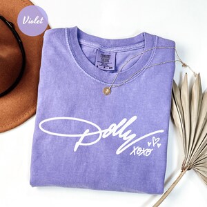 May include: A violet short-sleeved t-shirt featuring the white script text Dolly XOXO with three small heart icons. The shirt has a crew neckline and a Comfort Colors brand label visible at the inner collar, indicating a size small.