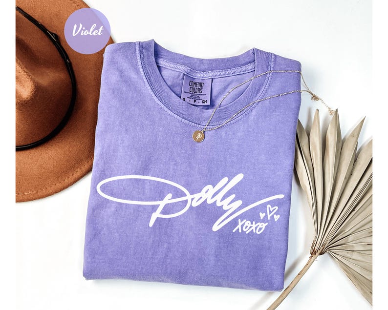 May include: A violet short-sleeved t-shirt featuring the white script text Dolly XOXO with three small heart icons. The shirt has a crew neckline and a Comfort Colors brand label visible at the inner collar, indicating a size small.
