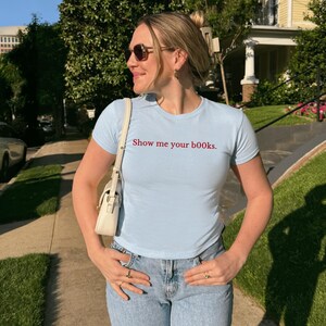 May include: A pale blue t-shirt with the text "Show me your books" in red. The shirt is worn with light blue jeans and a white handbag. The person is wearing sunglasses and rings.