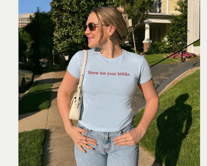 May include: A pale blue t-shirt with the text "Show me your books" in red. The shirt is worn with light blue jeans and a white handbag. The person is wearing sunglasses and rings.